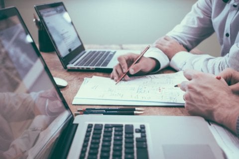 2 people sitting a table with laptops, looking at a sheet of notes