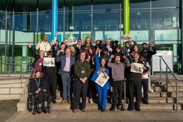 Group of staff. students and employers outside the college building cheering and celebrating, holing newspapers